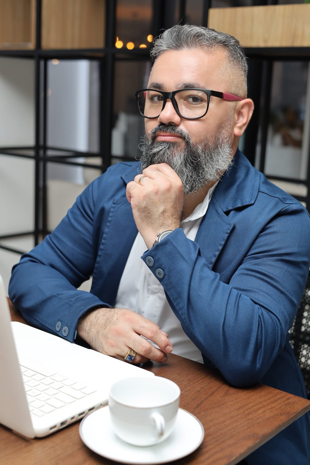 Thiago Diniz sentado à mesa de trabalho, com blazer azul, em atitude reflexiva durante um atendimento online.